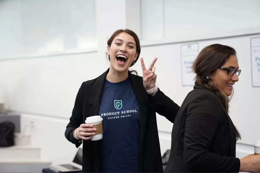 Two women smile and hold coffee cups while organizing a fundraiser for their school. One makes a peace sign with her fingers.